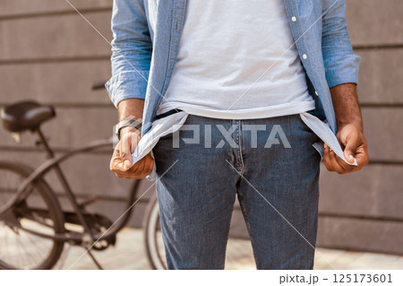 Poverty and absence of money. Unrecognizable afro man demonstrating empty turned out pockets outdoors, standing next to his bike, cropped image 125173601