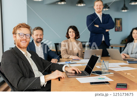 A team of business professionals is gathered around a large conference table, collaborating and working on projects. One man, with a red beard and glasses, is smiling and working on a laptop A team of business professionals is gathered around a large conference table, collaborating and working on projects. One man, with a red beard and glasses, is smiling and working on a laptop 125173675