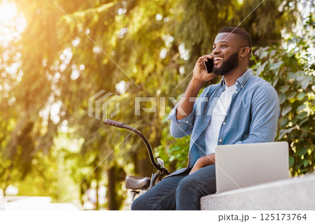 Cell Communication. Portrait Of Happy Handsome Afro Man Talking On Cellphone While Sitting With Laptop And Bicycle In Park Cell Communication. Portrait Of Happy Handsome Afro Man Talking On Cellphone While Sitting With Laptop And Bicycle In Park 125173764