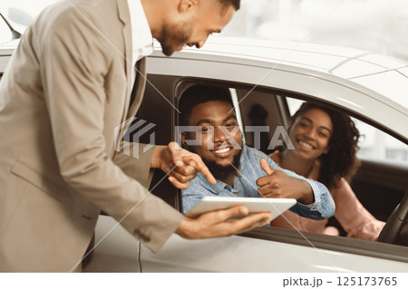 Buying New Car. Happy Black Couple Gesturing Thumbs Up Talking With Seller Approving New Automobile In Dealership Store Buying New Car. Happy Black Couple Gesturing Thumbs Up Talking With Seller Approving New Automobile In Dealership Store 125173765