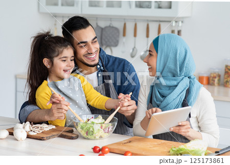 Happy Islamic Family Of Three Checking Food Recipe On Digital Tablet While Cooking In Kitchen Together, Cheerful Muslim Mom, Dad And Little Daughter Enjoying Preparing Lunch At Home, Closeup 125173861
