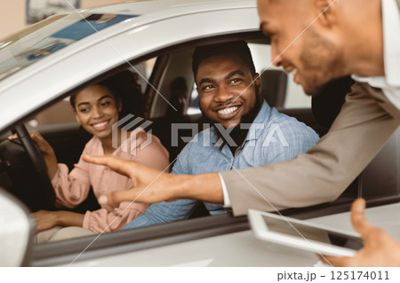 Joyful African Couple Buying New Car Talking With Salesman Testing Auto In Dealership Store. Selective Focus 125174011