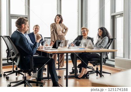 A group of business professionals are having a meeting in a modern office. The meeting is taking place around a large, rectangular table with several laptops and papers on it. 125174188