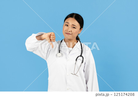 A healthcare professional Asian lady in a white coat expresses disapproval with a thumbs down gesture. She has a stethoscope around her neck, set against a bright blue background. A healthcare professional Asian lady in a white coat expresses disapproval with a thumbs down gesture. She has a stethoscope around her neck, set against a bright blue background. 125174208