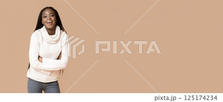A young black woman stands in a relaxed pose, arms crossed and smiling brightly against a warm, neutral backdrop. She showcases her long braided hair and casual sweater, copy space A young black woman stands in a relaxed pose, arms crossed and smiling brightly against a warm, neutral backdrop. She showcases her long braided hair and casual sweater, copy space 125174234