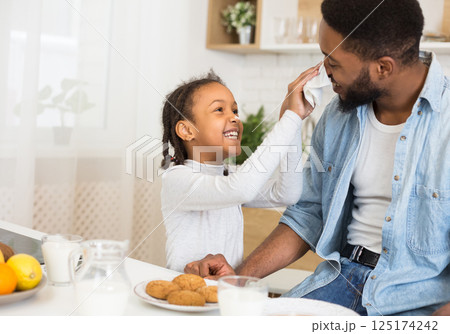 Afro Man And His Daughter Having Fun At Breakfast, girl cleaning father nouse with tissue, kitchen interior, copy space Afro Man And His Daughter Having Fun At Breakfast, girl cleaning father nouse with tissue, kitchen interior, copy space 125174242