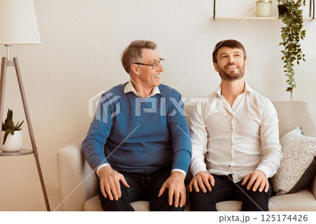 Cheerful Adult Son And Senior Father Talking And Smiling Sitting On Sofa At Home. Relationship With Elderly Parents. Selective Focus 125174426