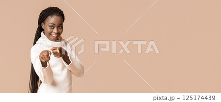 Attractive african woman pointing her index fingers at camera cheerfully, beige background, panorama with copy space 125174439