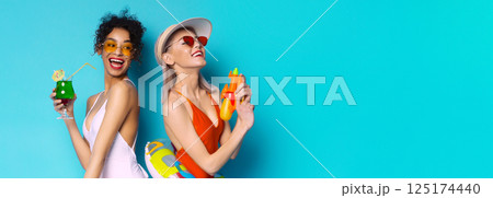 Two women in swimsuits are having fun in the summer sun. One woman is holding a cocktail and smiling, while the other woman is holding a water gun and laughing. The background is bright blue 125174440