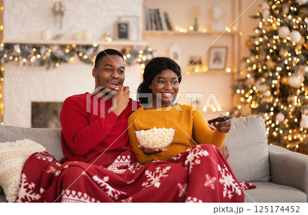 Free time, favorite movie and self-isolation at home. Millennial funny african american husband and wife with remote control and popcorn covered with blanket and watching tv in interior with tree Free time, favorite movie and self-isolation at home. Millennial funny african american husband and wife with remote control and popcorn covered with blanket and watching tv in interior with tree 125174452
