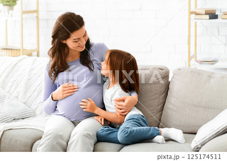 Pregnant woman and little daughter sitting on sofa at living room and having conversation, free space 125174511