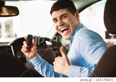 Happy Man Showing New Car Key To Camera Gesturing Thumbs Up Sitting In Auto In Dealership Store 125174625
