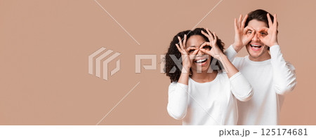 Joyful interracial couple fooling together, holding fingers near eyes, making funny glasses with ok signs, having fun over yellow background Joyful interracial couple fooling together, holding fingers near eyes, making funny glasses with ok signs, having fun over yellow background 125174681