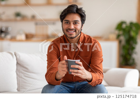 Arab man in an orange shirt sits on a white couch in a living room, smiling and holding a dark gray mug. He appears to be enjoying his coffee break. 125174718