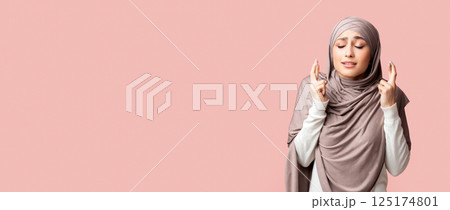 Young muslim girl in hijab crossing fingers, making wish, praying for miracle over pink studio background with free space 125174801