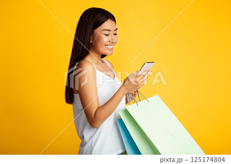 A smiling young Asian woman in a white tank top stands against a yellow background. She is holding a smartphone in her right hand and shopping bags in her left A smiling young Asian woman in a white tank top stands against a yellow background. She is holding a smartphone in her right hand and shopping bags in her left 125174804