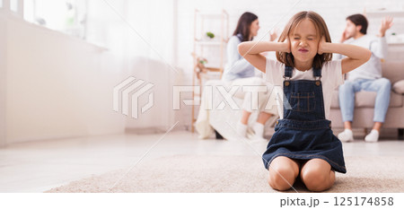 Little girl sitting on floor and covering ears with hands, tired of mom and dad arguing, quarrelling parents on background, selective focus on child 125174858