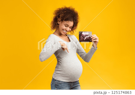Look here. Excited afro pregnant woman with ultrasound scan pointing at tummy over pink background Look here. Excited afro pregnant woman with ultrasound scan pointing at tummy over pink background 125174863