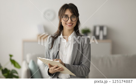 Happy professional middle eastern female psychologist writing in clipboard, looking and smiling at camera, working in modern office. Psychotherapy services, mental health professional concept 125174887