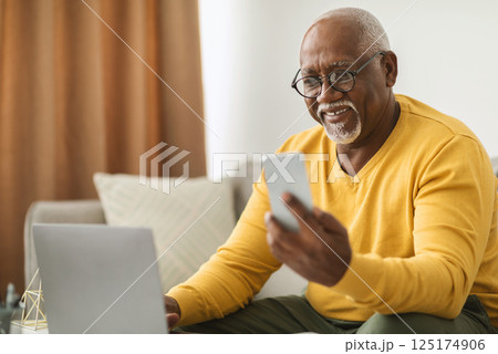 Senior Black Man Using Smartphone And Laptop Computer Working Online Sitting On Sofa Indoors. Senior Male Professional Texting Via Cellphone And Browsing Internet In Modern Office. Selective Focus 125174906