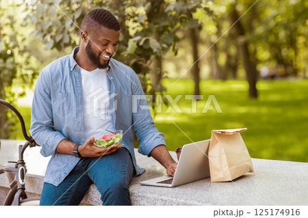 Happy afro businessman using laptop and having healthy lunch outdoors with bicycle parked nearby 125174916