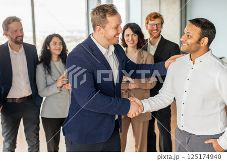 A group of diverse business professionals stand together in a modern office setting. A smiling man in a blue suit extends his hand to shake the hand of a new colleague, who also smiles warmly A group of diverse business professionals stand together in a modern office setting. A smiling man in a blue suit extends his hand to shake the hand of a new colleague, who also smiles warmly 125174940