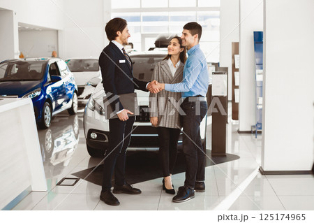 Successful Deal. Car seller handshaking with happy auto buyer couple standing in dealership store, selective focus 125174965