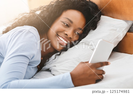 Close up of smiling black girl reading messages on smartphone in bed, having beautiful morning 125174981