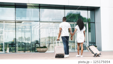 African American couple walking away from the entrance of an airport, each pulling a wheeled suitcase behind them. They are holding hands and walking away from the camera, copy space African American couple walking away from the entrance of an airport, each pulling a wheeled suitcase behind them. They are holding hands and walking away from the camera, copy space 125175054