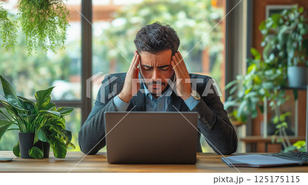 Stressed businessman feeling exhausted while working at a desk surrounded by plants 125175115