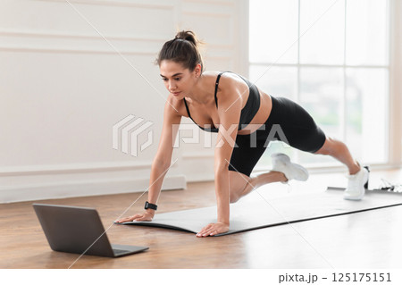 Online Training Concept. Focused sporty lady doing mountain climbers exercise, watching video tutorial or having online class on computer, looking at screen, exercising in her spatious apartment Online Training Concept. Focused sporty lady doing mountain climbers exercise, watching video tutorial or having online class on computer, looking at screen, exercising in her spatious apartment 125175151