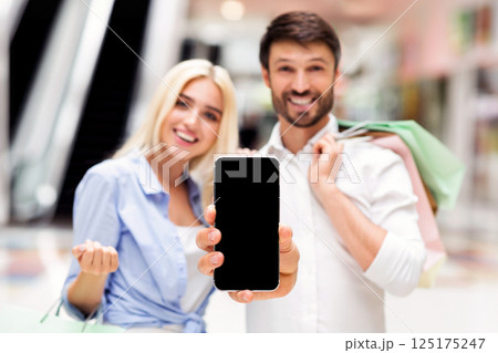 A man and woman, both smiling, stand in a shopping mall with bags, holding up a smartphone with black empty screen 125175247