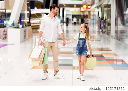 A smiling father and his daughter walk hand-in-hand through a brightly lit mall, each carrying shopping bags. They appear to be enjoying their time together and are excited about their purchases A smiling father and his daughter walk hand-in-hand through a brightly lit mall, each carrying shopping bags. They appear to be enjoying their time together and are excited about their purchases 125175252