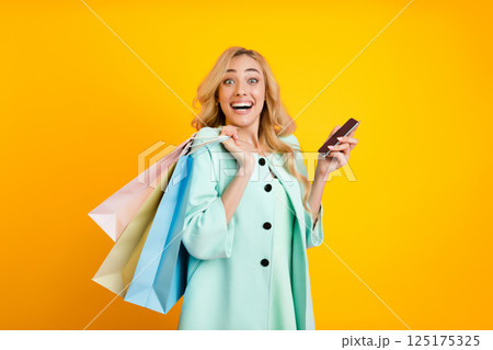 A young woman with long blonde hair is standing against a yellow background, holding a smartphone in her right hand and shopping bags in her left. She is looking at the phone 125175325