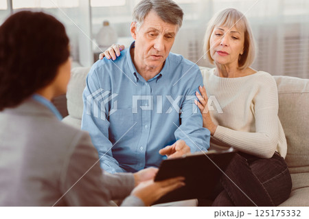 Last Will And Testament. Senior man sitting on couch with wife, checking document, signing papers with consultant 125175332