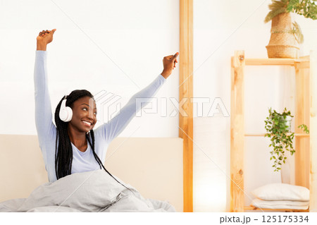 Good Morning. Happy Afro Lady In Headphones Stretching Arms Sitting In Cozy Bed At Home. Free Space 125175334