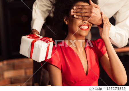 Romantic Surprise. Unrecognizable Afro Guy Giving Valentine Gift To Girlfriend Covering Her Eyes Celebrating In Restaurant. Cropped 125175383