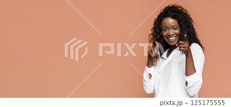 African American smiling woman with curly hair, wearing a white shirt, pointing her index fingers towards the camera. She is against a peach-colored background, and her expression is cheerful 125175555