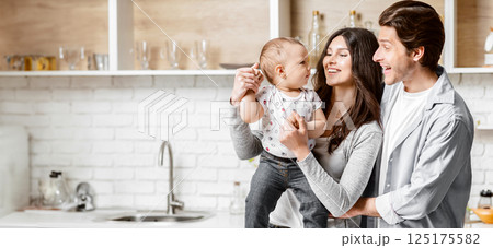 A young family of three stands in their modern kitchen, filled with white cabinets and brickwork. The mother holds their baby while the father looks on, beaming with joy, copy space 125175582