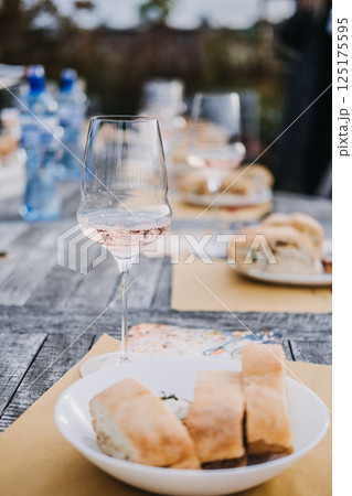 Glass of sparkling rose wine and a white bowl of rustic bread on a sunlit outdoor table with blurred guests in the background. Artisanal wine, grounded luxury, minimal menu, outdoor hospitality 125175595