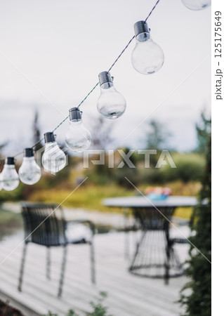 String of clear outdoor bulbs hanging above a garden patio with modern furniture in soft focus. Ambient garden lighting, outdoor entertaining, minimalist backyard design, cozy atmosphere 125175649