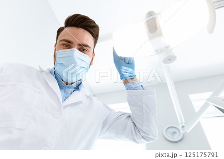 Male dentist in medical face mask turning on lamp, greeting patient in dental chair, looking at camera, copy space. Young man stomatologist beginning treatment, turning on light, closeup Male dentist in medical face mask turning on lamp, greeting patient in dental chair, looking at camera, copy space. Young man stomatologist beginning treatment, turning on light, closeup 125175791