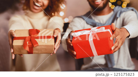 Excited afro couple holding xmas gift boxes and stretching them to camera in front of Christmas tree 125175823