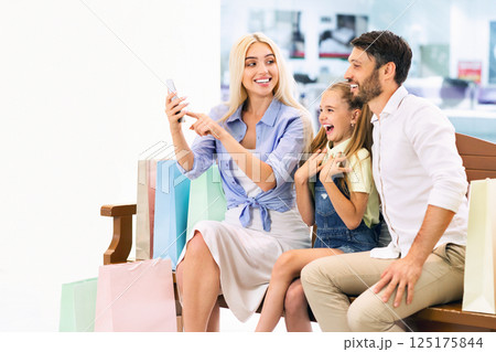 A family of three is sitting on a bench in a shopping mall, surrounded by shopping bags. The mother is holding a smartphone and taking a selfie, while the father and daughter are smiling 125175844
