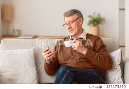 Retirement Lifestyle. Happy Senior Man Using Smartphone And Having Coffee Sitting On Couch Indoor. Selective Focus Retirement Lifestyle. Happy Senior Man Using Smartphone And Having Coffee Sitting On Couch Indoor. Selective Focus 125175850