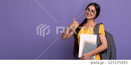 Happy indian female student wearing glasses showing thumbs up sign gesture, holding college materials and wearing backpack, standing isolated on purple studio background with free copy space, banner 125175855