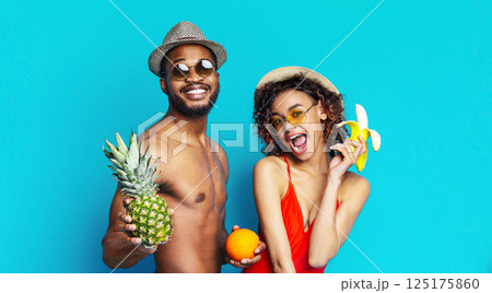 Black man and woman are smiling and posing for the camera against a blue background. The man is wearing sunglasses and a hat and holding a pineapple. The woman is holding a banana and an orange. Black man and woman are smiling and posing for the camera against a blue background. The man is wearing sunglasses and a hat and holding a pineapple. The woman is holding a banana and an orange. 125175860