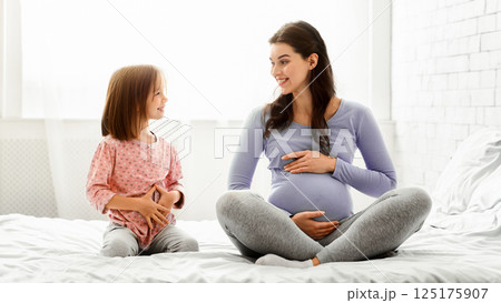 Expecting mother telling little girl about pregnancy, pregnant mother and preschool daughter touching bellies, sitting on bed Expecting mother telling little girl about pregnancy, pregnant mother and preschool daughter touching bellies, sitting on bed 125175907