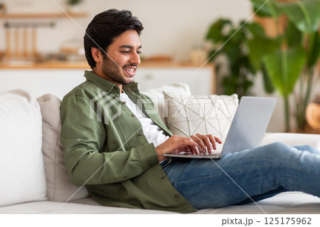 Arab man is sitting on a couch inside a home. He is smiling and using a laptop, appearing relaxed and comfortable. The man is wearing casual clothing, including a green shirt and blue jeans. Arab man is sitting on a couch inside a home. He is smiling and using a laptop, appearing relaxed and comfortable. The man is wearing casual clothing, including a green shirt and blue jeans. 125175962