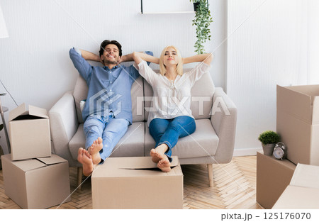 A relaxed couple sits on a couch in their new home, surrounded by cardboard boxes. 125176070